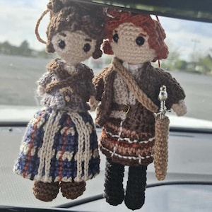 May include: Two handmade crochet dolls, one with a blue and white patterned skirt and the other with a brown outfit and a sword. Both dolls have dark hair and are hanging from a string. The dolls are likely decorative items or ornaments.