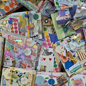May include: A collection of assorted craft ephemera packs featuring colourful paper scraps, stickers, ribbons, playing cards, and vintage postage stamps. Each packet is tied with string or ribbon, showcasing a variety of textures, patterns, and printed text.