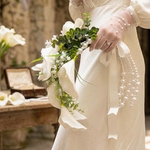 Könnte beinhalten: Ein Brautstrauß mit weißen Calla-Lilien, Grün und Perlen. Die Person trägt ein weißes Kleid und transparente Handschuhe. Der Text auf dem Bild lautet "CALLA LILY PEARL WATERFALL BOUQUET."