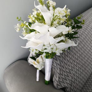 Peut inclure: Un bouquet de mariée et une boutonnière avec des callas blanches et de petites fleurs blanches. Le bouquet est enveloppé d'un ruban de satin blanc. Les tiges et le feuillage verts ajoutent du contraste. La boutonnière a un seul calla.