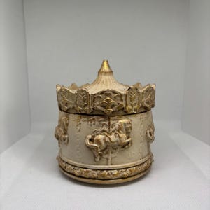 May include: A cream-coloured carousel-themed trinket box with gold accents. The box features embossed carousel horses and decorative trim. The lid is topped with a gold finial. The box is round and sits on a white surface.