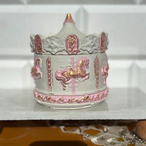 May include: A carousel-themed trinket box in white, pink, and gold. The box features carousel horses and decorative elements in pink with gold detailing. The lid is conical, and the base is round. The trinket box is a charming decorative item.