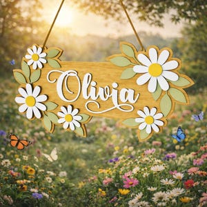 Peut inclure: Panneau en bois avec le nom "Olivia" en écriture blanche, orné de marguerites blanches à cœur jaune et de feuilles vertes. Le panneau est suspendu par une ficelle marron, avec des papillons en arrière-plan.