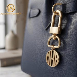 Pode incluir: Um charm de bolsa de metal dourado com um pingente de monograma redondo, preso a uma bolsa de couro azul marinho. O pingente apresenta as iniciais "MRB" em uma fonte estilizada. O charm é preso com um fecho e um anel. O texto "Personal MonogramCo" está no canto superior esquerdo.
