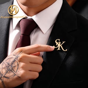 May include: A gold-colored monogram pin with the letters "SK" is pinned to a black blazer. The pin is being pointed at by a person's finger. The person is wearing a white collared shirt and a burgundy tie. The brand name "Personal Monogram Co" is visible.