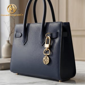 May include: A navy blue leather handbag with gold-coloured hardware. The bag has a top handle, a flap closure, and a decorative gold charm with initials. The brand name "Personal MonogramCo" is visible in the upper left corner.