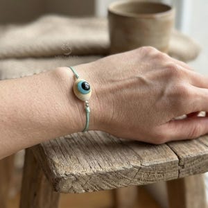 May include: A light blue cord bracelet featuring a wooden eye charm with a black pupil and light blue iris. The charm is set on a light wood base. The bracelet is displayed on a wooden surface.