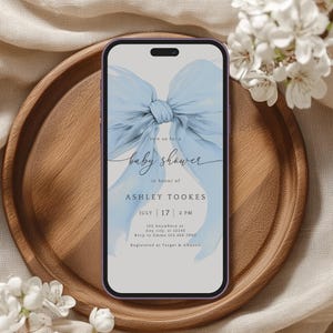 May include: A smartphone shows a baby shower invitation featuring a light blue bow. The invitation reads "Join us for a baby shower in honour of Ashley Tookes, July 17, 2 PM." The phone is on a wooden tray.