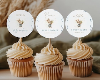 May include: Three cupcakes with light brown frosting, each topped with a round paper decoration. The decorations feature a watercolor illustration of a bunny holding flowers, with text that reads "Hello baby andrew", "Canner's BABY SHOWER", and "a little SWEET BUNNY".