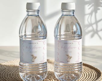 以下が含まれることがあります： 白いキャップが付いた透明なプラスチック製のウォーターボトルが2本。各ボトルには「Amelia's baby shower」というテキストと、花束を持ったウサギの水彩イラストが描かれたラベルが付いています。ボトルは織りマットの上にあります。