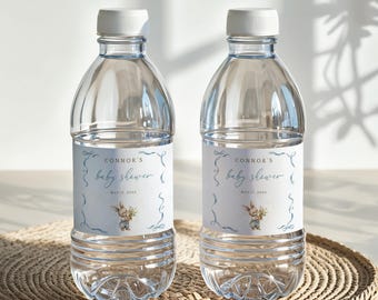 以下が含まれることがあります： 白いキャップが付いた透明なプラスチック製のウォーターボトルが2本。各ボトルには「Connor's baby shower」と書かれたラベルがあり、ウサギのイラストと2023年5月17日の日付が記載されています。ボトルは織り込まれた表面に置かれています。