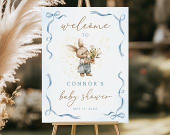 May include: A baby shower welcome sign on a gold easel. The sign features a watercolor illustration of a bunny holding flowers, with the text "welcome to Connor's baby shower" in elegant script, surrounded by blue ribbon.