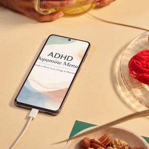 Op de afbeelding: Een smartphone met de tekst "ADHD Dopamine Menu" is aangesloten op een oplader. Een glas thee, een bord met een rood gebakje en een kom met gemengde noten zijn ook zichtbaar op een lichtgele ondergrond.