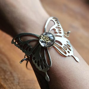 May include: A silver-toned butterfly bracelet with intricate cut-out details. The center features a clear, round element with visible gears. The bracelet is designed to be worn on the wrist, showcasing a unique steampunk aesthetic.