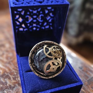 May include: A unique ring featuring a dark stone center with brass gears and a silver-toned outer band. The ring is displayed in an open, royal blue velvet ring box with a decorative top. A steampunk-inspired jewelry piece.