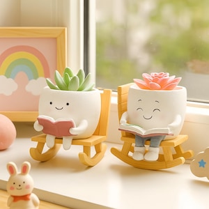 May include: Two white ceramic planters shaped like smiling faces, each holding a book and seated in a yellow rocking chair. One planter holds a green succulent, the other a pink flower. A whimsical home decor item.