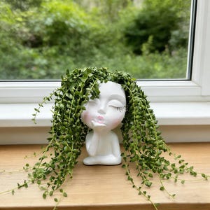 May include: A white ceramic planter shaped like a woman's face, with closed eyes and a kiss. The planter is filled with cascading green string of pearls plants, resembling hair. The planter sits on a wooden surface near a window.
