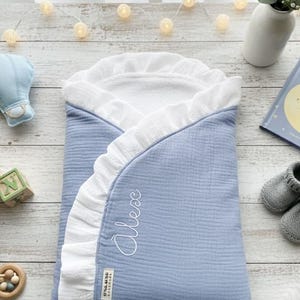 Puede incluir: Una manta envolvente para bebé en azul claro con un ribete blanco con volantes. La manta tiene el nombre "Alex" bordado en cursiva. La manta está hecha de una tela suave y texturizada. Una pequeña etiqueta con texto es visible.