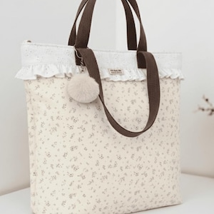 Puede incluir: Bolso tote crema con estampado floral y asas marrones. El bolso presenta un ribete de encaje blanco y un charm de pompón esponjoso. El bolso tiene forma rectangular y tiene cierre de cremallera.