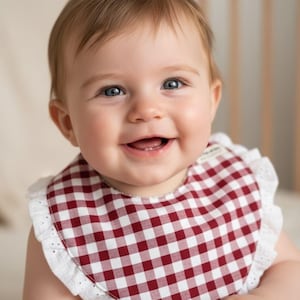 May include: A smiling baby wearing a red and white checkered bib with a white lace trim. The bib is around the baby's neck. The baby has light brown hair and blue eyes.