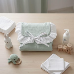 Puede incluir: Un cambiador de bebé verde claro con un borde blanco con volantes y un lazo blanco. El cambiador está rodeado de artículos esenciales para bebés, incluyendo un conejito de cerámica blanco, un sonajero de tortuga de ganchillo y paños blancos.