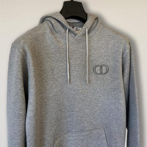 May include: A grey hoodie with a drawstring hood and a kangaroo pocket. The hoodie has a black and silver logo on the chest. The garment is hanging on a black hanger against a white wall.