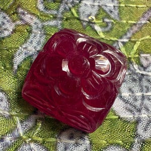 May include: A square-shaped, deep red gemstone bead featuring a carved floral design on its top surface. The translucent material displays a polished finish with intricate petal details etched into the center and corners of the square form.
