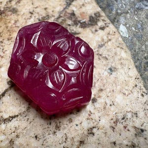 May include: A carved, deep red gemstone with a floral design. The gemstone is a rich, saturated color and features a central flower surrounded by detailed leaf patterns. The stone is set against a speckled, light-colored surface.