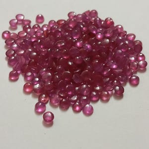 May include: A collection of small, polished, ruby-coloured gemstones. The gems are round or oval, with a translucent quality, and are scattered across a light surface. The stones vary in shade from light pink to deep red.