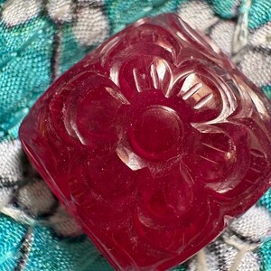 以下が含まれることがあります： 表面に花の彫刻が施された、半透明の深い赤色の正方形のビーズ。ビーズは角が丸く磨き上げられた仕上げで、中央の円形要素を6枚の彫刻された花びらが囲むデザインが特徴です。