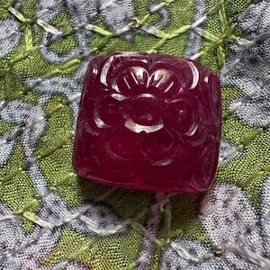 以下が含まれることがあります： 深いマゼンタ色の正方形の宝石ビーズで、上面に花の彫刻が施されています。石は半透明で、滑らかで丸みを帯びたエッジと磨かれた仕上げが特徴であり、素材に刻まれた花びらの複雑なディテールがはっきりと見て取れます。