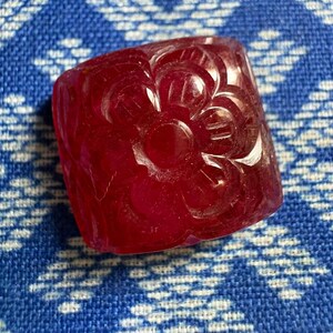 Könnte beinhalten: Ein quadratischer, tiefroter Edelstein-Bead mit einem geschnitzten Blumenmuster auf der Oberfläche. Die Gravur zeigt einen zentralen runden Punkt, der von sechs einzelnen Blütenblättern umgeben ist, was ein strukturiertes Relief auf dem Stein erzeugt.