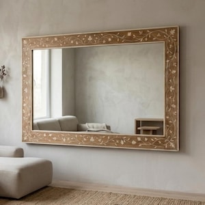 May include: A large rectangular mirror with a light brown wooden frame. The frame features a delicate floral and vine pattern in a lighter shade. The mirror is mounted on a neutral-toned wall.