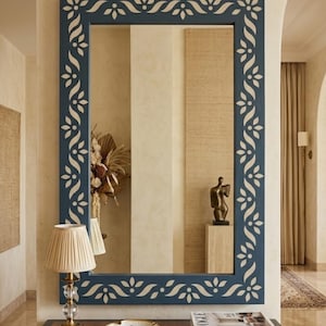 May include: A large rectangular mirror with a navy blue frame and a white floral design. The mirror hangs above a dark wood table with a lamp, a decorative plate, and books. The overall style is elegant and decorative, suitable for home decor.