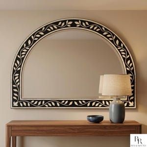 May include: An arched mirror with a black frame and white leaf pattern. The mirror is above a wooden console table. A grey lamp with a beige shade and a blue bowl are on the table. The mirror is a decorative home decor item.