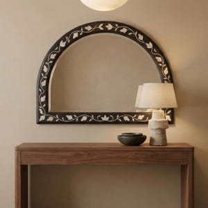 May include: A wooden console table with an arched mirror above. The mirror has a black frame with white floral detailing. A lamp with a beige shade and a bowl sit on the table. A white light fixture hangs above.
