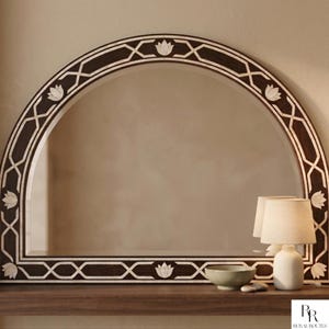May include: An arched mirror with a dark brown frame, decorated with a white geometric and floral pattern. The mirror is above a dark wooden surface, with a small lamp and bowl reflected in the glass.