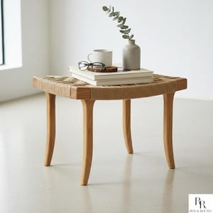 Puede incluir: Un pequeño taburete cuadrado de madera con una parte superior tejida. Las patas son curvas y de madera clara. Una taza, libros, gafas y un jarrón con vegetación están encima. El taburete mide aproximadamente 46 cm de ancho y 46 cm de largo.