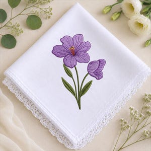 May include: A square white fabric napkin featuring an embroidered purple flower design with green stems and leaves. The napkin is finished with a delicate white lace trim along the edges, creating a clean and detailed textile accessory for table settings.