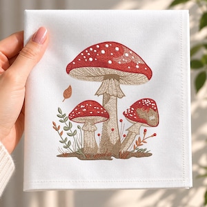 Peut inclure: Une serviette de table carrée blanche ornée d'une broderie détaillée représentant trois champignons rouges à pois blancs. Le champignon central est le plus grand, entouré de deux plus petits, de tiges feuillues vertes et d'une feuille orange.