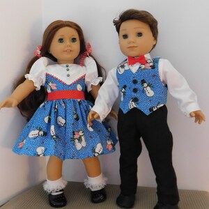 May include: Two dolls dressed in snowman-themed outfits. The girl doll wears a blue dress with a red belt and white ruffled sleeves. The boy doll wears a white shirt, blue vest with snowman pattern, black pants, and a red bow tie.