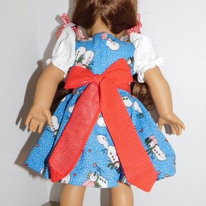 May include: A doll wearing a blue dress with a red bow and a snowman pattern. The dress has white puffed sleeves with red trim. The doll has long brown hair and is wearing white socks with lace trim and black shoes.