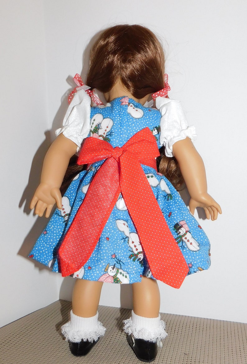 May include: A doll wearing a blue dress with a red bow and a snowman pattern. The dress has white puffed sleeves with red trim. The doll has long brown hair and is wearing white socks with lace trim and black shoes.