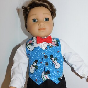 May include: A doll wearing a white shirt, a blue vest with a snowman pattern, and a red bow tie.