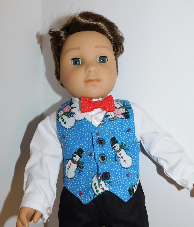 May include: A doll wearing a white shirt, a blue vest with a snowman pattern, and a red bow tie.