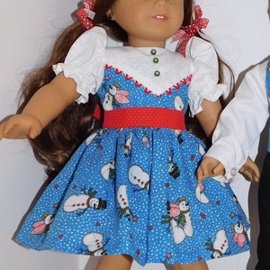 May include: A doll wearing a blue dress with a snowman pattern, a red belt, and white socks. The doll has long brown hair and blue eyes.