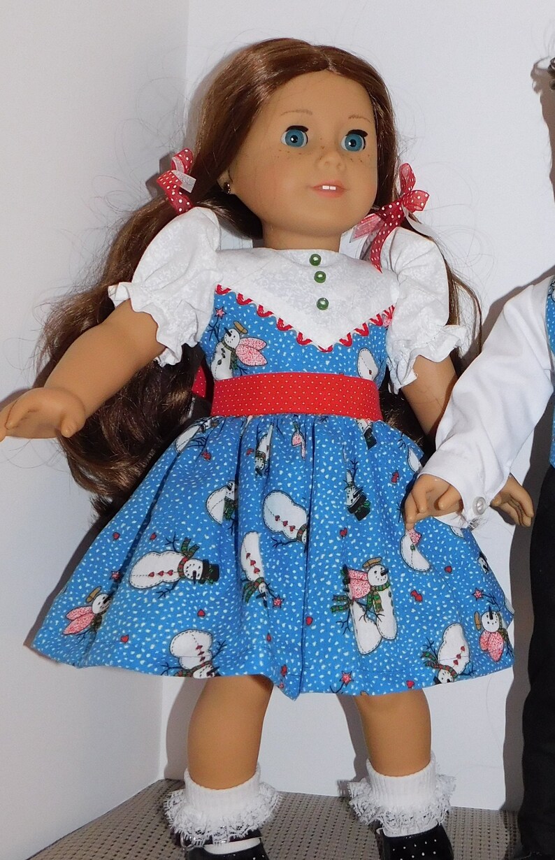 May include: A doll wearing a blue dress with a snowman pattern, a red belt, and white socks. The doll has long brown hair and blue eyes.