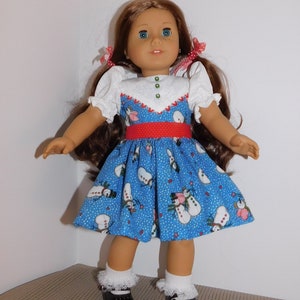 May include: A doll wearing a blue dress with a snowman pattern, a red belt, and white puffed sleeves. The doll has brown hair, blue eyes, and is wearing black shoes with white socks.