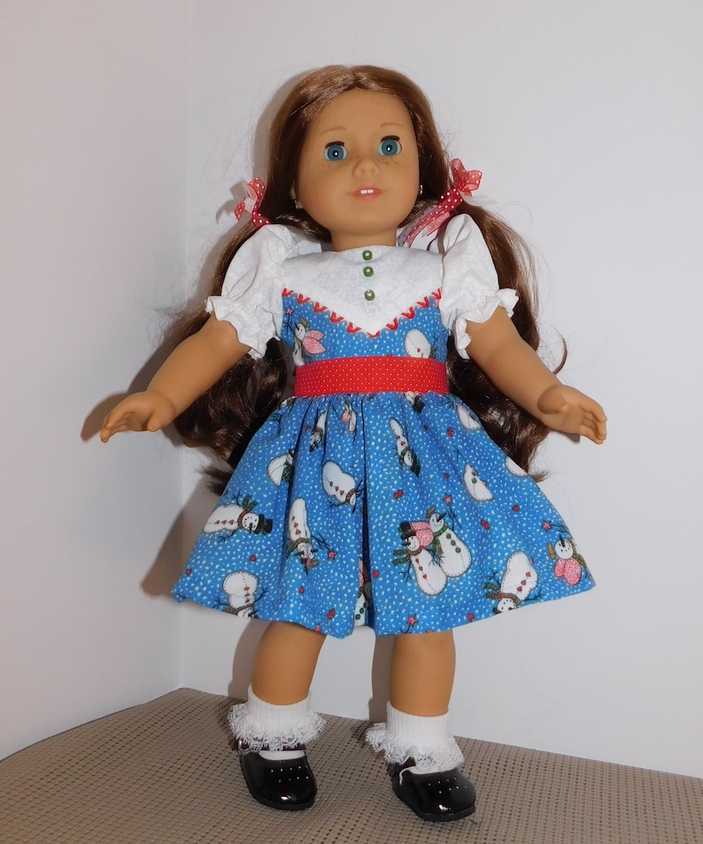 May include: A doll wearing a blue dress with a snowman pattern, a red belt, and white puffed sleeves. The doll has brown hair, blue eyes, and is wearing black shoes with white socks.