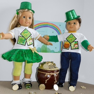 May include: Two dolls dressed in St. Patrick's Day outfits. One doll wears a green skirt, yellow leggings, and a white t-shirt with a shamrock design. The other doll wears blue jeans and a white t-shirt with a pot of gold design. Both dolls wear green hats.